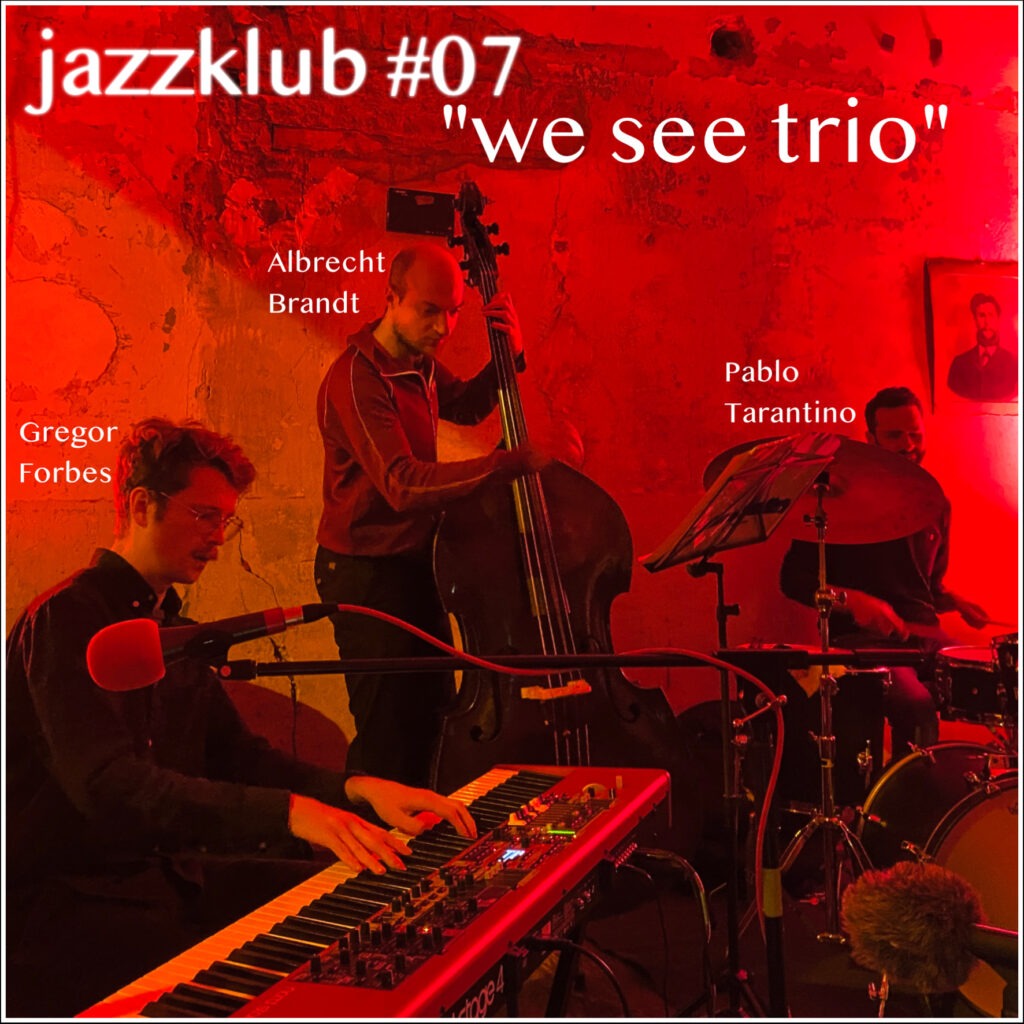 Ein Episodenbild. In einem rot beleuchteten Raum spielt ein Jazztrio, bestehend aus einem Pianisten an seinem E-Piano (links), einem Kontrabassisten (Mitte) und einem Schlagzeuger (rechts). Vor dem Pianisten ragt ein Mikrofon mit einem roten Windschutz ins Bild (ganz links). Rechts über dem Schlagzeuger hängt ein sehr altes Schwarz-Weiß-Photo eines bärtigen Mannes an der Wand (ganz rechts).